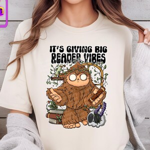 May include: A white t-shirt with a cartoon illustration of a Bigfoot holding a book. The text "It's giving big reader vibes" is printed on the shirt.