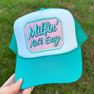 May include: A teal and white trucker hat with the text "Milfin' Ain't Easy" embroidered on a pink patch. The hat has a curved brim and a mesh back. The Rainbow Emporium Gifts logo is visible in the top left corner.