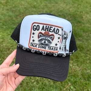 May include: Black and white trucker hat with a patch featuring a raccoon wearing a cowboy hat and the text "GO AHEAD... TALK SOME TRASH". The hat has a black brim and mesh back.