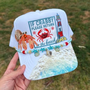 May include: White trucker hat with a beach-themed design. The hat features a crab and lighthouse graphic, the text "IF CRABBY PLEASE RETURN to the beach", and a beaded accent. The brim has a beach scene.