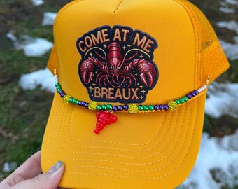 May include: Mustard yellow trucker hat with a mesh back and a curved brim. The hat features a patch with a red crawfish and the text "COME AT ME BREAUX". A beaded necklace with a red crawfish charm is attached to the brim.