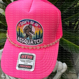 May include: A pink trucker hat with a white patch that says "Don't be an Assquatch" with a cartoon Bigfoot in the center. The hat has a beaded trim and a white label that says "Otto World Famous Foam Front OSFM Trucker Durable Snap Closure".