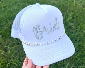 May include: White trucker hat with the word "Bride" in rhinestones and a pearl and letter charm that reads "Miss to Mrs."