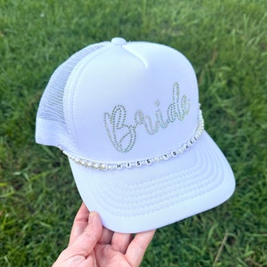 May include: White trucker hat with the word "Bride" in rhinestones and a pearl and letter charm that reads "Miss to Mrs."