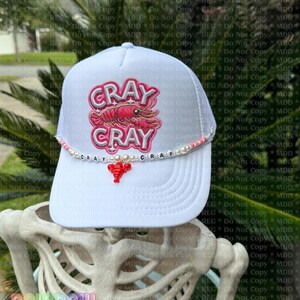 May include: White trucker hat with a mesh back and a curved brim. The front features the words "CRAY CRAY" in pink and red, with a lobster graphic. A beaded accent and lobster charm add detail.