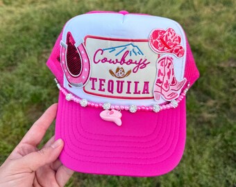 May include: Pink and white trucker hat with a pink cowboy boot, horseshoe, and mountain patch. The patch says "Cowboys & Tequila". The hat has a pink brim and a white mesh back. The hat is decorated with a pink and white beaded trim.