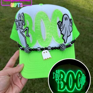 May include: A neon green and white Halloween-themed trucker hat with the word "BOO" in large, neon green letters. The hat features a skeleton and ghost applique, and a beaded accent. The hat is held by a person.