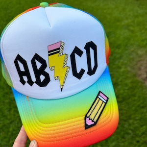 May include: A white and rainbow trucker hat with the text "ABCD" in black gothic font and a yellow lightning bolt with a pink eraser and leopard print. A yellow pencil with a pink eraser is on the side of the hat.