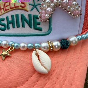 May include: A coral-colored trucker hat with a white front panel featuring the text "HELLS SHINE" in teal. The hat is adorned with a beaded necklace, a seashell, and a gold starfish charm. A large, pearl-studded starfish decorates the top.