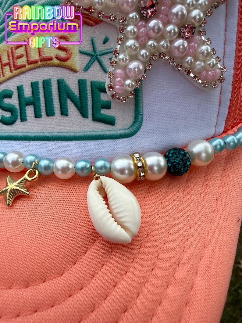 May include: A coral-colored trucker hat with a white front panel featuring the text "HELLS SHINE" in teal. The hat is adorned with a beaded necklace, a seashell, and a gold starfish charm. A large, pearl-studded starfish decorates the top.