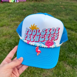 May include: A blue and white trucker hat with the text "Poolside Princess" in pink and white letters. The hat has a white brim with a row of white pearls and a pink flamingo charm.