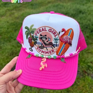 May include: Pink trucker hat with a white front panel featuring an embroidered patch that says "Feral Girl Summer" with a raccoon, palm trees, and a flamingo. The hat has a pink mesh back and a pink brim with a beaded trim and a pink flamingo charm.