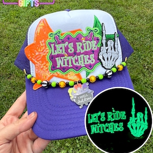 May include: A purple and white trucker hat with the text "Let's Ride Witches" and a skeleton hand. The hat features an orange sequined star and a beaded accent. A small charm reads "I smell children".