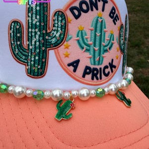 May include: A white trucker hat with a coral bill and a beaded accent. The hat features a sequined cactus patch and a pink patch with a cactus and the words "DON'T BE A PRICK". A green cactus charm hangs from the beaded accent.