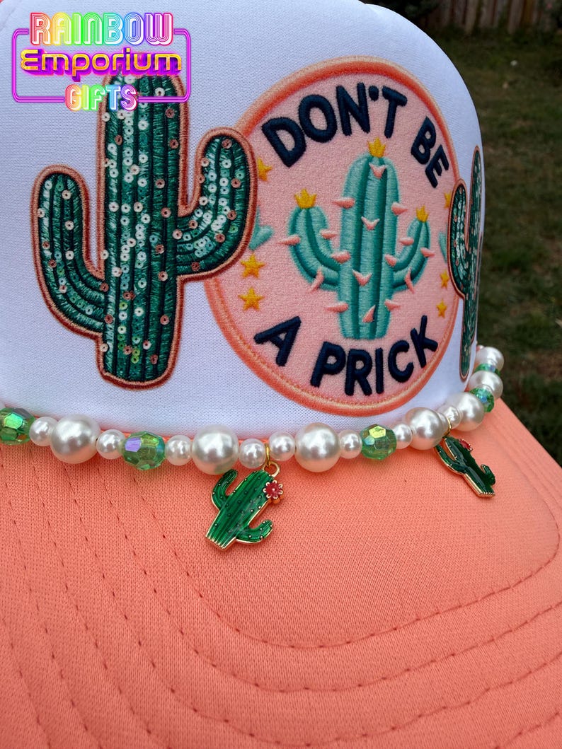 May include: A white trucker hat with a coral bill and a beaded accent. The hat features a sequined cactus patch and a pink patch with a cactus and the words "DON'T BE A PRICK". A green cactus charm hangs from the beaded accent.