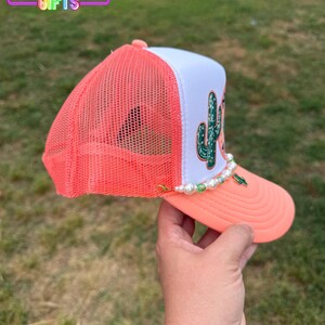 May include: A coral and white trucker hat with a cactus design. The hat features a coral mesh back, a white front panel with a green cactus graphic, and a coral brim. A pearl and green bead chain decorates the brim. The text "Rainbow Emporium Gifts" is visible.