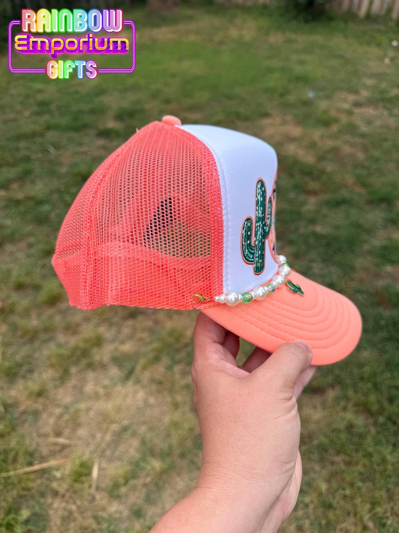 May include: A coral and white trucker hat with a cactus design. The hat features a coral mesh back, a white front panel with a green cactus graphic, and a coral brim. A pearl and green bead chain decorates the brim. The text "Rainbow Emporium Gifts" is visible.