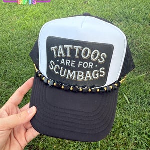 May include: Black and white trucker hat with the text "TATTOOS ARE FOR SCUMBAGS" on a patch. The hat has a mesh back, a curved brim, and is decorated with black and gold star-shaped embellishments.