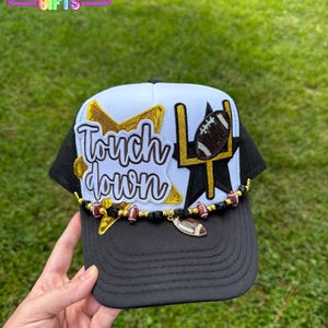 May include: Black and white trucker hat with a football theme. The hat features the words "Touchdown" in gold script, a football, and a goalpost design. A beaded chain with football charms adorns the brim. The hat is decorated with gold stars.