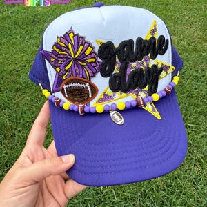 May include: Purple and white baseball cap with the text "Game Day" in black. The cap is decorated with a football, a pom-pom, and star-shaped accents. The brim is purple, and the top is white with purple mesh.