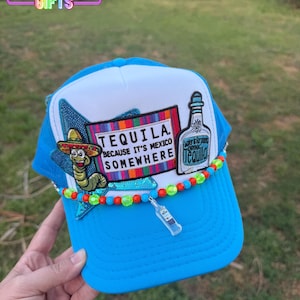 May include: A blue and white trucker hat with a tequila theme. The hat features a patch with the words "Tequila because it's Mexico somewhere" and a bottle of tequila. It is decorated with colorful beads and a small tequila bottle charm.