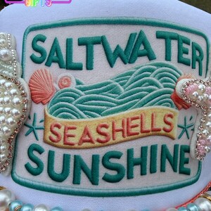 May include: A white hat with a green and white embroidered patch that reads "Saltwater, Seashells, Sunshine." The patch features a wave design and seashell accents. The hat is decorated with pearls and seashells.