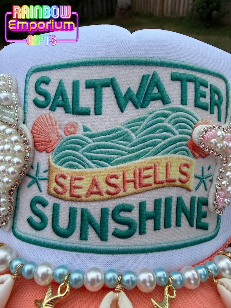 May include: A white hat with a green and white embroidered patch that reads "Saltwater, Seashells, Sunshine." The patch features a wave design and seashell accents. The hat is decorated with pearls and seashells.