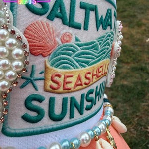 May include: A white hat with the words "SALTWATER, SEASHELLS, SUNSHINE" embroidered in green and gold. The hat features a seashell and wave design. A pearl and seashell necklace is draped over the hat.
