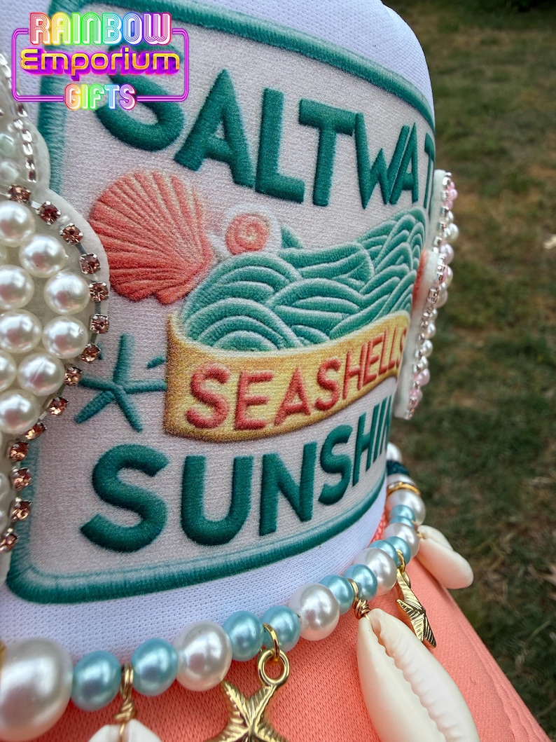 May include: A white hat with the words "SALTWATER, SEASHELLS, SUNSHINE" embroidered in green and gold. The hat features a seashell and wave design. A pearl and seashell necklace is draped over the hat.