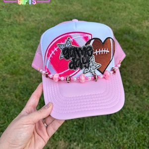 May include: Pink and white trucker hat with a football theme. The hat features a pink football, a brown heart-shaped football, and the words "Game Day". A beaded chain adorns the brim. The hat is displayed on a green grassy background.