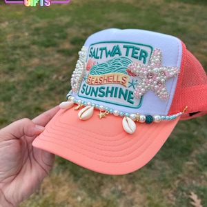 May include: A coral and white trucker hat with a mesh back. The hat features a beaded design with seashells, pearls, and a starfish. The front of the hat has the words "Saltwater, Seashells, Sunshine".