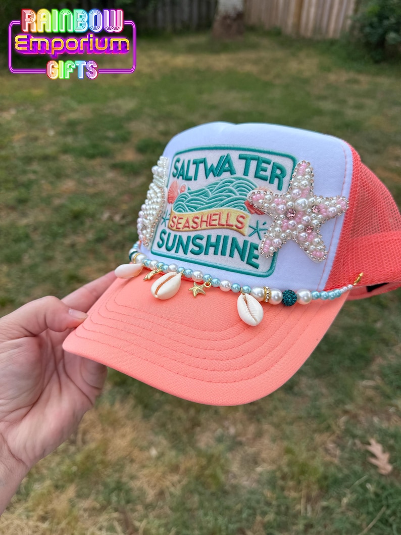 May include: A coral and white trucker hat with a mesh back. The hat features a beaded design with seashells, pearls, and a starfish. The front of the hat has the words "Saltwater, Seashells, Sunshine".