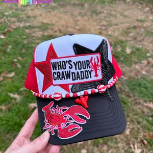 May include: A black and red trucker hat with the text "WHO'S YOUR CRAW DADDY" and a red lobster graphic. The hat features a red star and black accents, with a beaded trim and a small lobster charm.