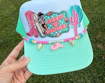 May include: A mint green and white trucker hat with a surf-themed design. The hat features a patch with a cowgirl on a surfboard, the text "Surf's Up Cowgirl", and decorative embellishments including a pink cowboy hat and seashells.