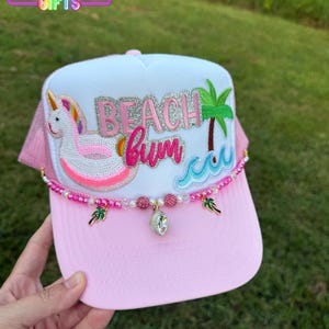 May include: Pink and white trucker hat with the text "Beach Bum" and a unicorn floatie design. The hat has a pink brim and a beaded embellishment with charms. The text "Rainbow Emporium Gifts" is at the top.