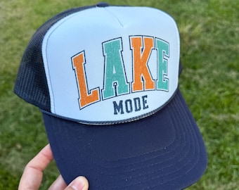 May include: A navy blue and white trucker hat with the words "LAKE MODE" in orange and teal lettering. The hat has a black mesh back and a curved brim. The text "RAINBOW Emporium GIFTS" is at the top.