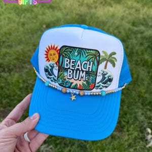 May include: A blue and white trucker hat with a beach scene and the text "Beach Bum". The hat has a gold beaded trim and a gold star charm.
