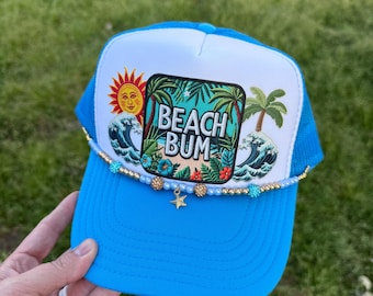 May include: A blue and white trucker hat with a beach scene and the text "Beach Bum". The hat has a gold beaded trim and a gold star charm.