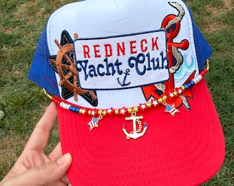 May include: A red, white, and blue trucker hat with the words "Redneck Yacht Club" embroidered on the front. The hat features a nautical theme with an anchor, ship's wheel, and star charms. The brim is red, the top is white, and the mesh is blue.