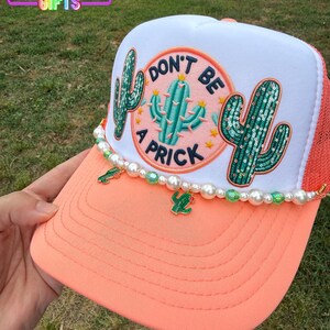 May include: A coral and white trucker hat with a cactus design and the phrase "DON'T BE A PRICK." The hat features a beaded trim with cactus charms. The brim is coral, and the mesh back is orange.
