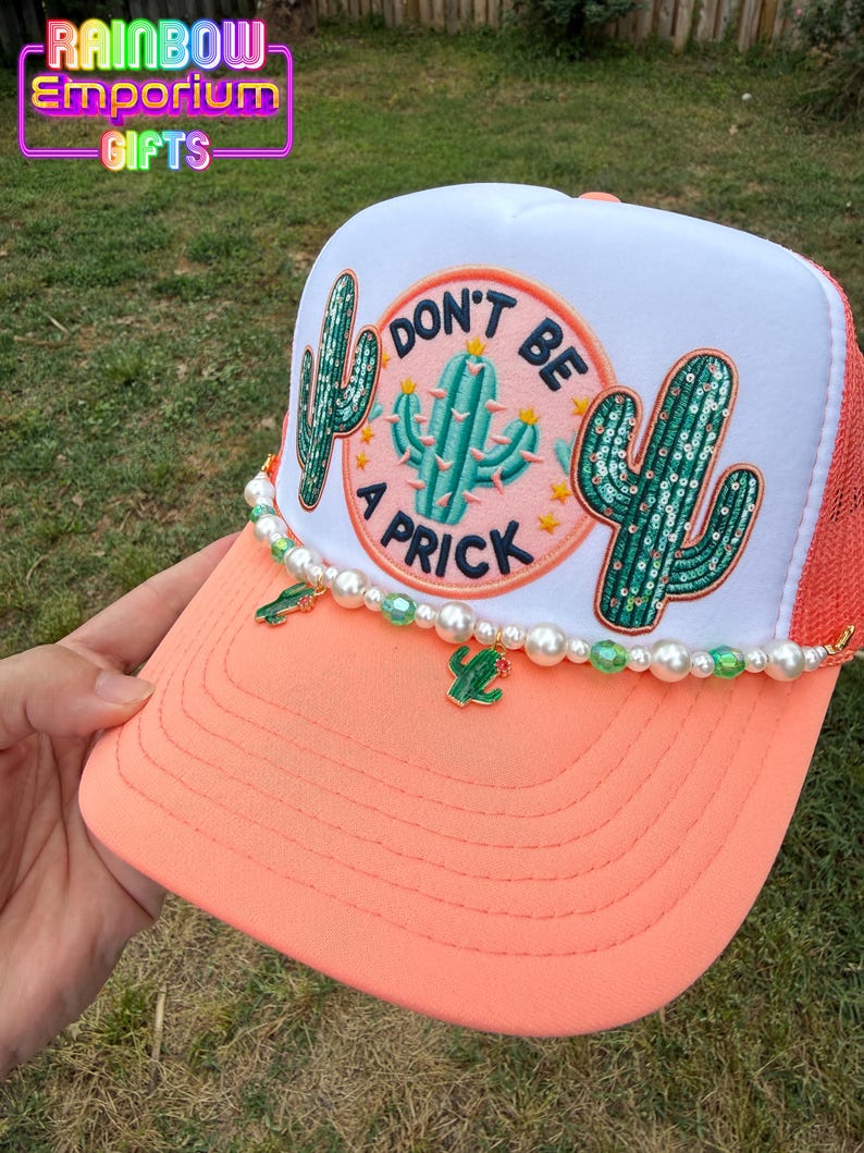 May include: A coral and white trucker hat with a cactus design and the phrase "DON'T BE A PRICK." The hat features a beaded trim with cactus charms. The brim is coral, and the mesh back is orange.
