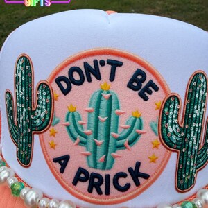 May include: A white trucker hat with a coral brim and a beaded accent. The hat features a circular patch with a teal cactus and the words "DON'T BE A PRICK". Two sequined cactus designs flank the central patch. The text "Rainbow Emporium Gifts" is at the top.