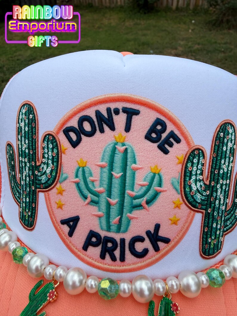 May include: A white trucker hat with a coral brim and a beaded accent. The hat features a circular patch with a teal cactus and the words "DON'T BE A PRICK". Two sequined cactus designs flank the central patch. The text "Rainbow Emporium Gifts" is at the top.
