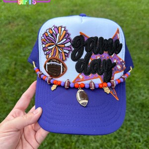 May include: Purple and white trucker hat with "Game Day" text and football-themed embellishments. Features a football charm, a beaded chain, and a pom-pom design. The hat has a purple brim and a white top.