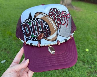 May include: A white and maroon trucker hat featuring an embroidered football, pom-poms, and a silver tag. The hat is accented with a beaded trim of white pearls, small footballs, and black beads, with a dangling football charm at the center.