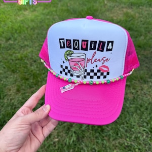 May include: A hot pink trucker hat with a white front panel featuring the words "Tequila please" and a cocktail graphic. The hat has a pink mesh back and a beaded accent. The brim is hot pink.