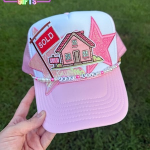 May include: A light pink and white trucker hat featuring a large pink glitter star patch, a house patch, and a red sold sign patch. A beaded string with white pearls, pink beads, and letter beads spelling out JUST SOLD is attached across the front.