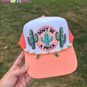 May include: A coral and white trucker hat with a cactus design. The hat features the text "DON'T BE A PRICK" in a circular design, with cactus illustrations. The brim is coral, and the hat is adorned with pearl and cactus charms.