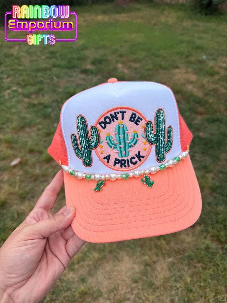 May include: A coral and white trucker hat with a cactus design. The hat features the text "DON'T BE A PRICK" in a circular design, with cactus illustrations. The brim is coral, and the hat is adorned with pearl and cactus charms.