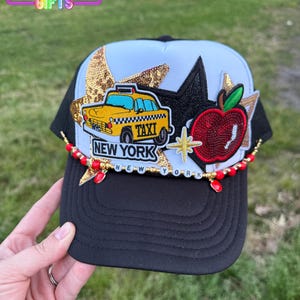 May include: A black and white trucker hat with a yellow taxi cab, red apple, and gold star patches. The hat has a black brim and a white mesh back. The hat also has a red and gold beaded trim with the words "NEW YORK" in gold letters.